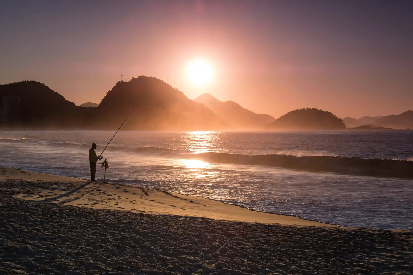 Silhouetted fisherman standing on the beach holding a long fishing rod against a vibrant sunset backdrop.